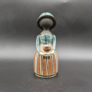 Puede incluir: Una figurita de cerámica de una mujer que lleva un vestido azul y verde y una falda a rayas marrones y blancas. Lleva un pequeño cuenco marrón en sus manos. La figurita está pintada en un estilo de arte popular.