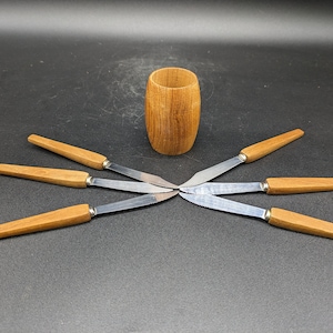 Könnte beinhalten: Ein Satz von fünf Buttermessern mit Holzgriffen und silbernen Klingen, die in Sternform um einen Holzbecher angeordnet sind.