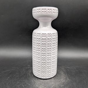 May include: A white ceramic vase with a cylindrical body and a flared, wide rim. The exterior surface features a repeating embossed grid pattern of small, square indentations covering both the main body and the upper rim section.
