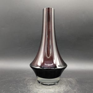 May include: A tall, elegant vase with a flared base and a slender neck. The vase has a dark, reflective finish that transitions from a deep purple to black at the base. The clear glass base adds a touch of transparency.