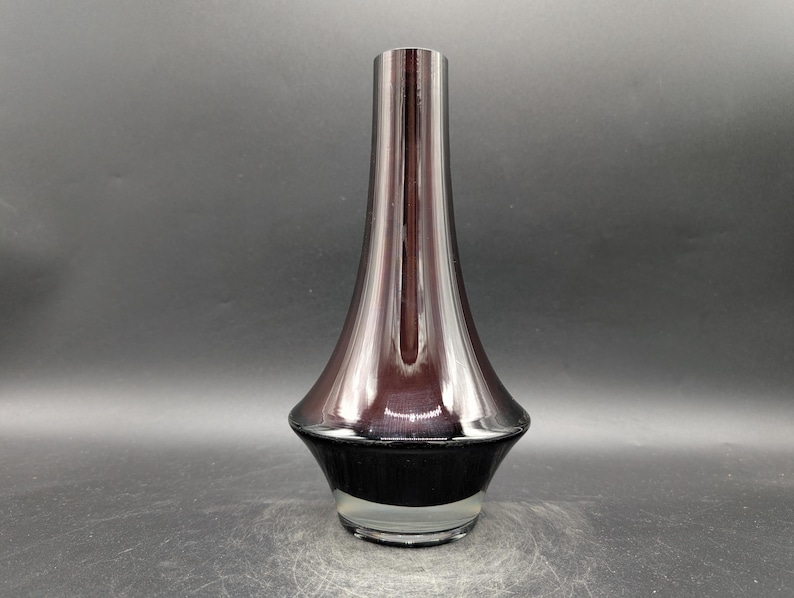 May include: A tall, elegant vase with a flared base and a slender neck. The vase has a dark, reflective finish that transitions from a deep purple to black at the base. The clear glass base adds a touch of transparency.