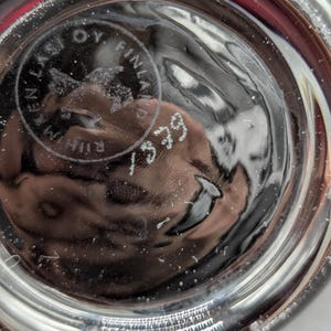 May include: Close-up of the base of a clear glass object. The base has a circular design with the text "EN LASI OY FINLAND" and the number "1379". The glass has a textured surface.