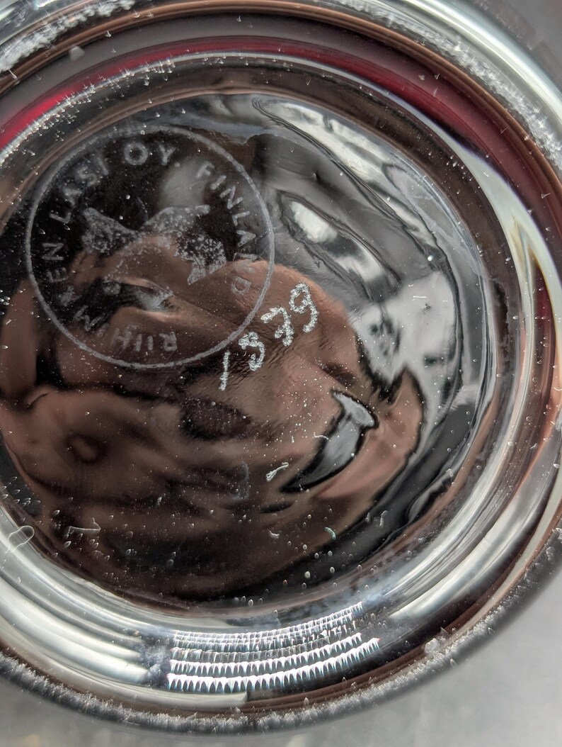 May include: Close-up of the base of a clear glass object. The base has a circular design with the text "EN LASI OY FINLAND" and the number "1379". The glass has a textured surface.