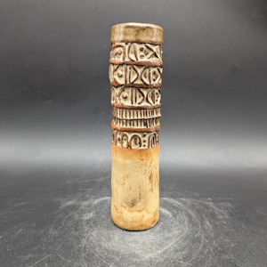 May include: A tall, cylindrical ceramic vase with a textured surface. The vase features bands of carved geometric patterns in shades of brown and cream. The base is a lighter tan color, and the top rim is a darker brown.