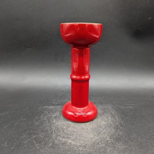 Puede incluir: Un portavelas de cerámica rojo brillante. Presenta una base circular, un tallo cilíndrico y una parte superior acampanada. El acabado brillante del portavelas refleja la luz, resaltando sus curvas suaves.