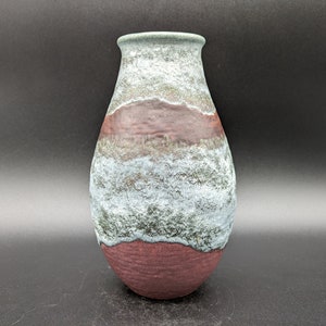 Puede incluir: Un jarrón de cerámica con un esmalte texturizado y moteado en tonos de verde, marrón y blanco. El jarrón tiene una forma redondeada y bulbosa y una abertura estrecha.