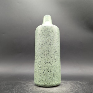 Könnte beinhalten: Eine hohe, grüne Flasche mit gesprenkelter Textur. Die Flasche hat einen zylindrischen Körper und einen schmalen Hals. Die Farbe ist ein gedämpftes Grün mit dunkleren grünen Sprenkeln, wodurch ein einzigartiges, strukturiertes Aussehen entsteht. Die Flasche ist wahrscheinlich aus Keramik oder Glas.