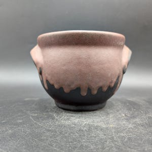 May include: A small, handcrafted ceramic bowl with a unique design. The upper portion is a muted pinkish-brown, transitioning to a dark, dripped black glaze at the base. Features two small handles on either side. The bowl sits on a dark surface.
