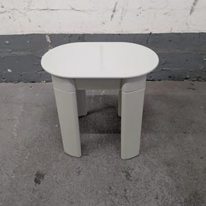 May include: A small, light gray side table with an oval top and four rectangular legs. The table is made of a solid material and is suitable for indoor use. The table is approximately 18 inches long, 12 inches wide, and 16 inches tall.