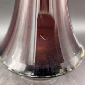 May include: Close-up of a clear, flared glass vase with a deep burgundy interior. The vase has a smooth, reflective surface with a slight imperfection. The base of the vase has a subtle, golden-yellow rim.
