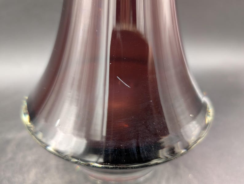 May include: Close-up of a clear, flared glass vase with a deep burgundy interior. The vase has a smooth, reflective surface with a slight imperfection. The base of the vase has a subtle, golden-yellow rim.