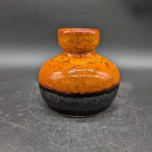 Könnte beinhalten: Kleine Keramikvase mit rundem Körper und schmalem Hals. Die Vase ist mit einem Farbverlauf glasiert, der von einem dunkel schwarzen Sockel zu einem leuchtenden Orange und Gelb an der Oberseite übergeht. Die Vase hat eine strukturierte Oberfläche.