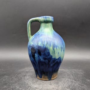 May include: A ceramic jug with a handle, showcasing a blend of blue and green glaze. The colours drip down the sides, creating a unique, artistic effect. The base is a natural, unglazed colour.