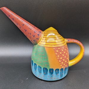 Könnte beinhalten: Eine farbenfrohe Gießkanne aus Keramik mit einem langen Ausguss und einem Griff. Die Kanne ist mit geometrischen Mustern in Rot-, Gelb-, Grün-, Blau- und Orangetönen verziert.
