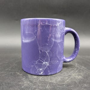 Könnte beinhalten: Eine lilafarbene Keramiktasse mit einem weißen Craquelé-Glaze. Die Tasse hat einen Henkel und ist am Rand leicht abgebrochen.