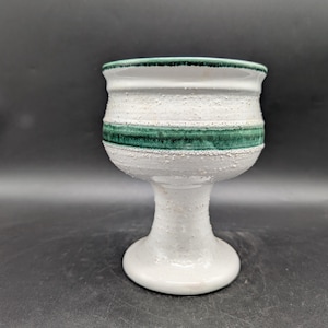 Könnte beinhalten: Ein weißer Keramikbecher mit strukturierter Oberfläche und einem grünen Band in der Mitte. Der Rand ist ebenfalls grün. Der Becher hat einen Sockel und steht vor einem dunklen Hintergrund.