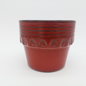 Puede incluir: Una maceta de cerámica roja con un borde decorativo. La maceta tiene un borde ligeramente acampanado y un acabado liso y brillante.