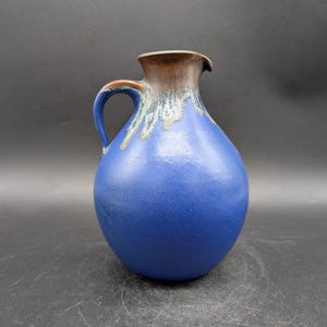 May include: A blue ceramic jug with a brown rim and handle. The jug has a rounded body and a curved handle. The rim has a drip effect with a light green colour. The jug is likely used for serving beverages.