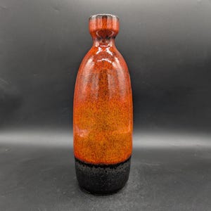 Könnte beinhalten: Eine hohe Keramikvase in Flaschenform. Die Vase hat einen schwarzen Sockel, einen orange-rot gesprenkelten Körper und einen dunkelbraunen, strukturierten Hals. Ein dekoratives Objekt.