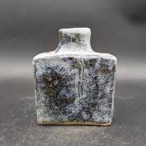 Könnte beinhalten: Kleine, quadratische Keramikflasche mit kurzem Hals. Die Flasche hat eine strukturierte Oberfläche mit einem gesprenkelten Muster aus Dunkelblau, Grau und Braun. Der obere Teil und der Hals sind hellgrau.