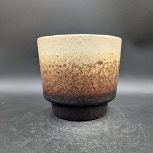 May include: A ceramic planter with a gradient color scheme. The top is a light beige, transitioning to brown, and ending with a dark brown base. The planter has a cylindrical shape with a slightly flared rim and a raised base.