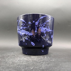 May include: A dark blue ceramic planter with a unique design. The planter features a speckled pattern of white and blue paint splatters across its surface. The planter has a cylindrical shape with a slightly flared base.