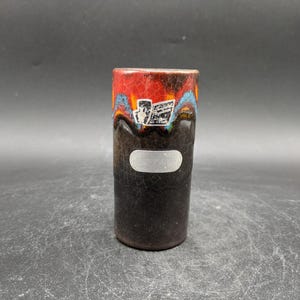 Może przedstawiać: Czarny ceramiczny wazon z czerwoną, pomarańczową i niebieską glazurą. Wazon ma białą etykietę z czarnym tekstem.