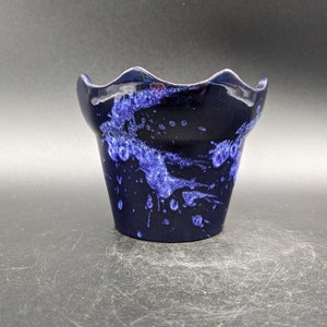 May include: A small, dark blue ceramic pot with a wavy rim and a speckled, abstract blue design. The pot has a cylindrical shape and sits on a dark surface. The interior is also dark blue.