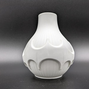 May include: A white ceramic vase with a bulbous body and a narrow neck. The vase features vertical ribbed detailing and a series of arched, decorative cutouts around the midsection. The vase is set against a dark background.