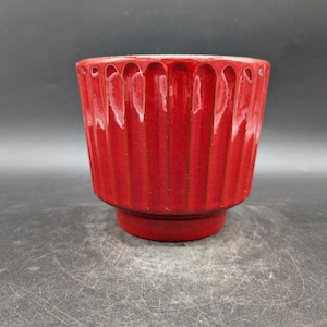 May include: A bright red ceramic plant pot with a ridged exterior design. The small, round pot has a pedestal base and a smooth interior. The glossy finish reflects light, highlighting the texture. Suitable for small plants or succulents.