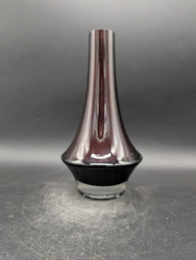 May include: A tall, elegant vase with a flared base and a slender neck. The vase has a gradient color scheme, transitioning from a dark, opaque base to a translucent, reddish-brown upper portion. The vase is made of glass and has a glossy finish.