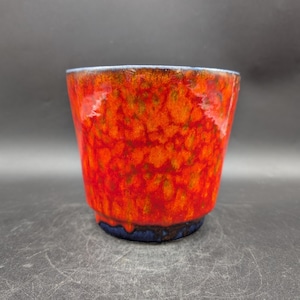 Può includere: Una tazza in ceramica con una vivace glassa strutturata nei toni del rosso, dell'arancione e con accenni di verde. Il bordo ha una sottile linea blu e la base è blu scuro. La tazza ha una forma leggermente svasata.