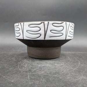 Könnte beinhalten: Eine Keramikschale mit einem weißen oberen Bereich mit einem schwarzen, abstrakten Design. Die Schale hat einen dunkelbraunen Sockel und einen passenden Rand. Das Design ist ein sich wiederholendes Muster aus geschwungenen Linien. Die Schale ist wahrscheinlich für dekorative oder funktionelle Zwecke.