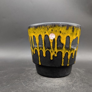 Könnte beinhalten: Ein schwarzer und gelber Keramik-Pflanztopf mit einem einzigartigen Tropfglasur-Design. Die gelbe Glasur scheint die Seiten des Topfes hinunterzutropfen. Der Topf hat eine runde Form und einen kleinen Aufkleber auf der Vorderseite.