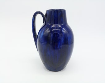 May include: A dark blue ceramic pitcher with a handle. The pitcher has a glossy finish with streaks of lighter blue, creating a marbled effect. The handle is curved and attached to the side of the pitcher. The pitcher is a decorative item.
