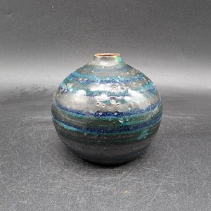 May include: A round ceramic vase with a dark gray base and horizontal bands of teal and blue. The vase has a small opening at the top and a glossy finish. The overall shape is spherical.