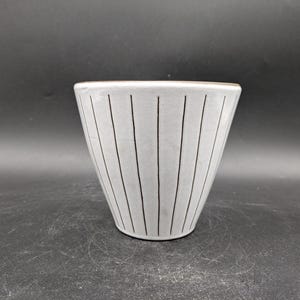 Op de afbeelding: Een witte keramische plantenpot met zwarte verticale strepen. De pot heeft een ronde bodem en een brede opening.