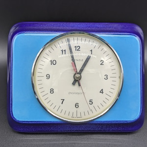 May include: A blue and purple square clock with a white face and black hands. The clock face has black numbers and the text "chronocquartz" and "FRENZLE" printed on it.