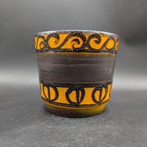 Könnte beinhalten: Eine zylindrische Keramikvase mit einem dunkelbraunen Band und orangefarbenen und schwarzen dekorativen Mustern. Ober- und Unterseite weisen ein sich wiederholendes Design auf, während das Mittelband schlicht ist. Die Vase steht auf einer dunklen Oberfläche.
