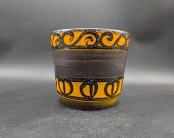 May include: A cylindrical ceramic vase with a dark brown band and orange and black decorative patterns. The top and bottom feature a repeating design, while the middle band is plain. The vase is on a dark surface.