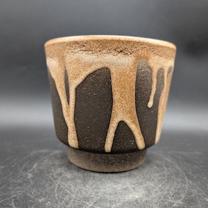 Peut inclure: Une tasse en céramique avec une base marron foncé et un effet de glaçure marron clair dégoulinant autour du bord. La tasse a une surface texturée et une petite base circulaire.