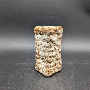 Könnte beinhalten: Kleine, rechteckige Keramikvase mit strukturiertem, geriffeltem Design. Die Vase ist überwiegend weiß mit braunen Akzenten und hat ein leicht raues, gealtertes Aussehen. Geeignet für kleine Blumen oder Dekorationszwecke.