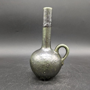 Könnte beinhalten: Eine kleine, dunkelgrüne Keramikvase mit strukturierter Oberfläche und einem Henkel. Die Vase hat einen schmalen Hals und einen runden Körper.