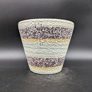 Pode incluir: Um vaso de cerâmica branco com listras marrons, amarelas e verdes. O vaso tem uma superfície texturizada e uma borda ligeiramente alargada.