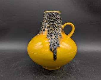 May include: A yellow ceramic vase with a rounded body and a small handle. The top section of the vase has a textured, dark speckled pattern. The vase has a glossy finish and is set against a dark background.