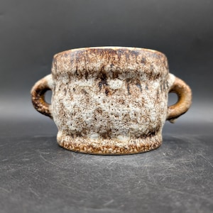 Könnte beinhalten: Eine Keramiktasse mit zwei Henkeln, mit einer strukturierten Oberfläche mit einer gesprenkelten weißen und braunen Glasur. Die Tasse hat eine abgerundete Form und einen dunkelbraunen Rand und Boden. Die Gesamtästhetik ist rustikal und erdig.