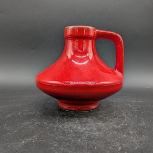 Könnte beinhalten: Eine rote Keramikvase mit rundem Korpus und einem einzigen Henkel. Die Vase hat eine glänzende Oberfläche.