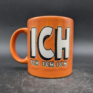 Könnte beinhalten: Orangefarbene Keramiktasse mit einem weißen Design, auf dem "ICH" und "ICH ICH ICH" in schwarzer Schrift stehen.