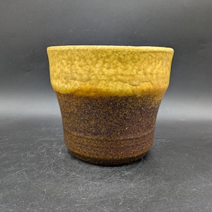 Könnte beinhalten: Eine Keramiktasse mit strukturierter Oberfläche. Die obere Hälfte ist hellgelbbraun, während die untere Hälfte dunkler braun ist. Die Tasse hat eine leicht ausgestellte Form.