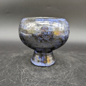 Könnte beinhalten: Eine Keramikschale mit rundem Körper und Sockel. Die Schale hat eine metallische Oberfläche mit Blau-, Silber- und Schwarztönen. Der Innenraum ist dunkel und der Rand hat einen blauen Farbton. Die Schale ist wahrscheinlich handgefertigt.